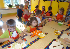 Grupa dzieci siedzi przy stole nakrytym podkładkami z rozłożonymi deskami, w ręku trzymają noże, którymi kroją owoce.